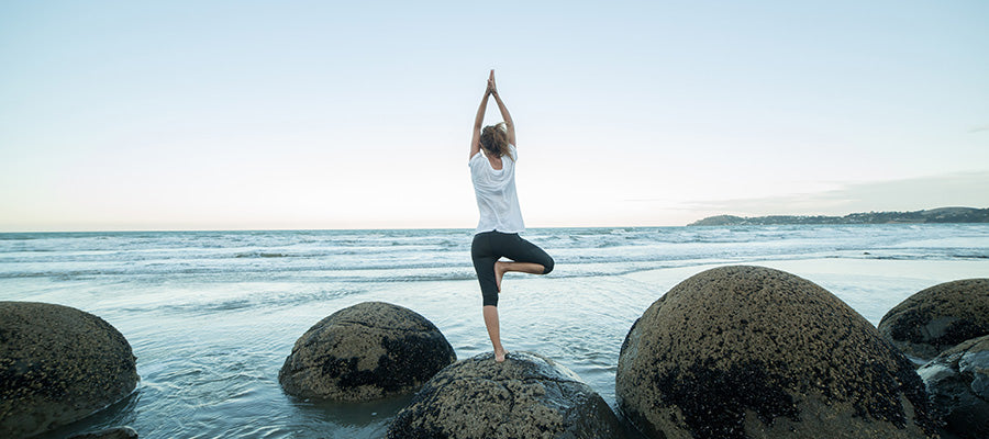 Woman doing yoga on a large rock on a beach facing the water.