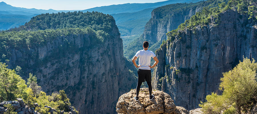Man standing on top of a rock over looking a ravine and other mountains.