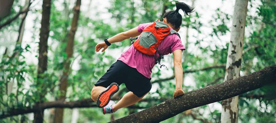 Athletic ponytailed woman with backpack doing parkour over a large tree limb through a green forest