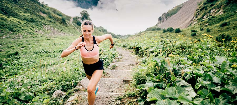 Athletic woman running up rock stairs on a mountain side surrounded by green plants with yellow flowers.