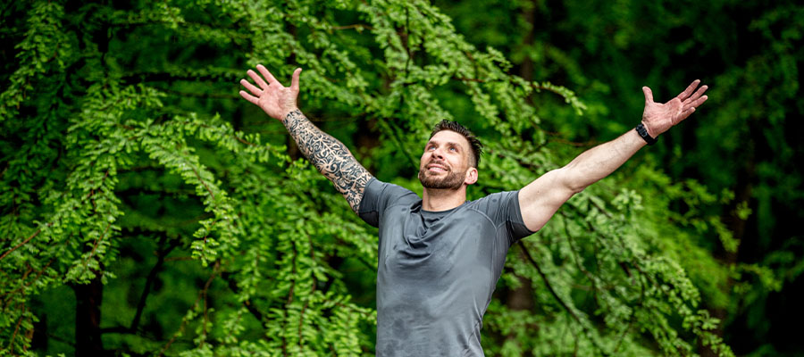 Man looking up to the sky with his arms held wide open, standing next to a large vibrant green bush.