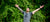 Man looking up to the sky with his arms held wide open, standing next to a large vibrant green bush.