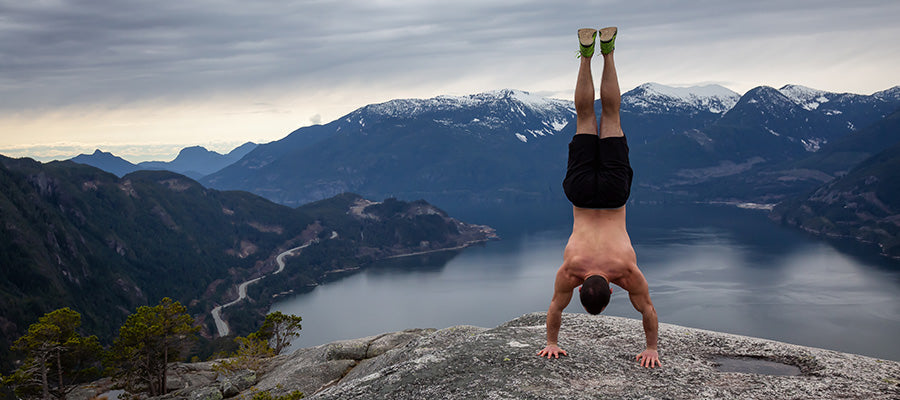 Athletic male doing a handstand on a cliffside above a mountain lake.