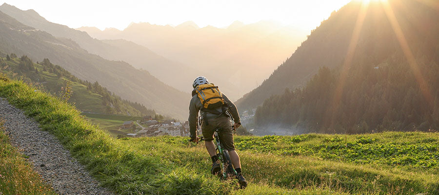 Person riding a mountain bike away down a grassy trail in the mountains with the sunset in the background