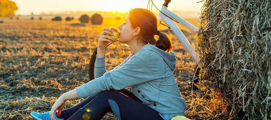 Athletic woman takes a break from a bike ride to snack on an apple while resting by a hay bale at sunset