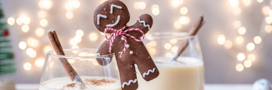 Gingerbread man standing next to two glasses of eggnog that have cinnamon sticks in them.