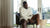 BAM Adebayo in a white shirt and shorts sitting on a chair by a window with a view of greenery messaging his calves.