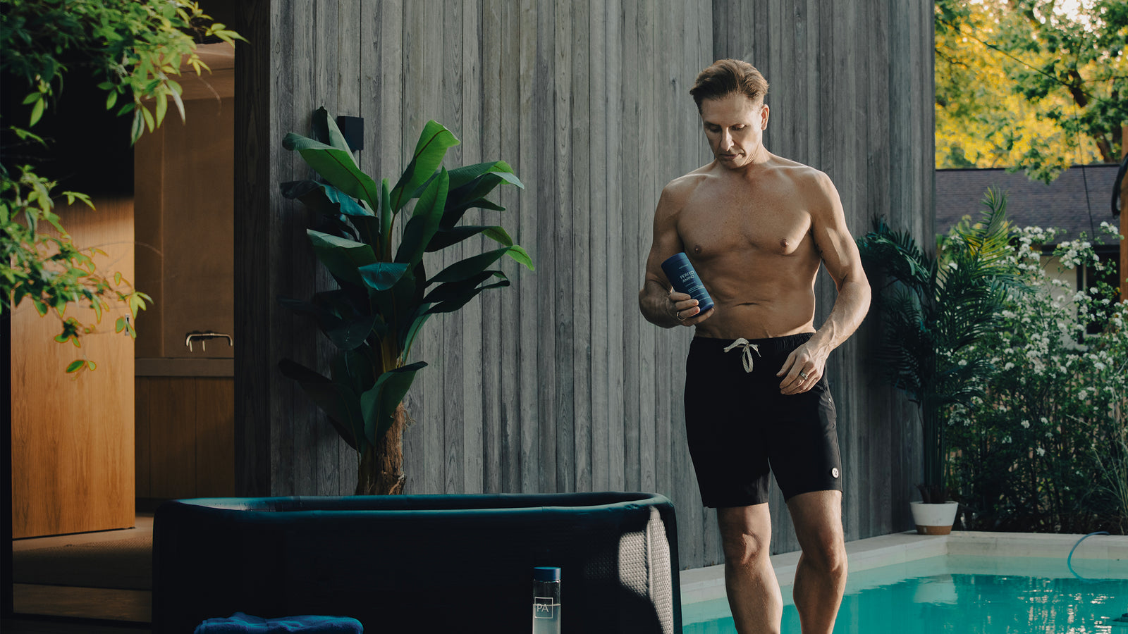 Dave Asprey in black swim shorts holding a blue bottle of Perfect Amino by a pool and cold plunge